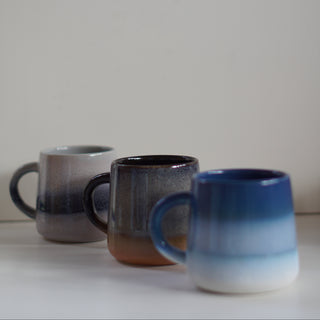 Three ceramic mugs with ombre gradients in gray, brown, and blue, arranged on a white surface against a plain light gray background.