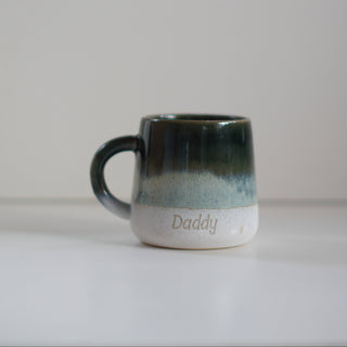 A small handmade ceramic mug with a dark green and blue glaze fading to a white base, featuring the word 'Daddy' printed on the front, set against a plain light gray background.