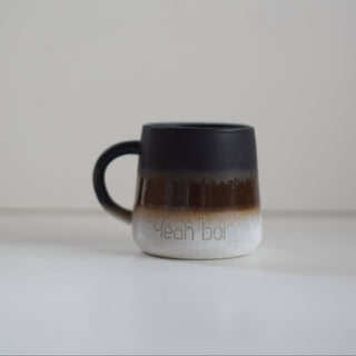 A ceramic coffee mug with a black-to-brown-to-white gradient glaze, featuring the text 'Yeah Boi' printed on the white section, set against a plain light gray background.