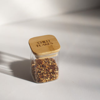 A small clear glass jar filled with red chili flakes, topped with a light bamboo lid engraved with 'CHILLI FLAKES', set on a white surface with dramatic diagonal shadows.