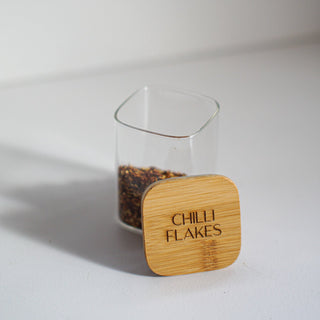 A small clear glass jar containing red chili flakes, with a square light wood lid placed in front. The lid is engraved with the words 'CHILLI FLAKES'.