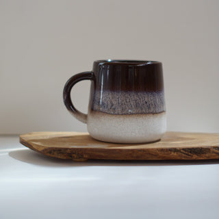 A handmade ceramic mug with a dark brown to white gradient glaze, resting on a rustic wooden slice against a neutral light gray background.