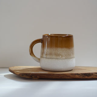 A ceramic mug with a brown and white gradient glaze, resting on a rustic wooden board against a neutral light gray background.