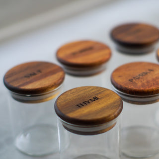 Clear glass spice jars with round wooden lids engraved with names including THYME, SALT, and PAPA CHOCOLATE, arranged on a light surface.