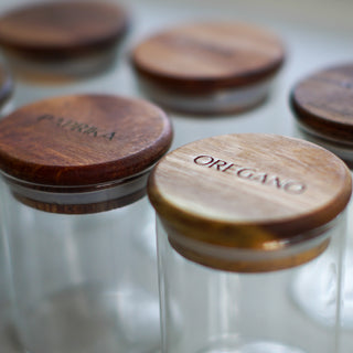 Clear glass jars with round wooden lids engraved with spices, including OREGANO and paprika, on a light neutral background.