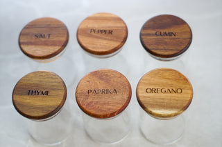 Six clear glass spice jars with round wooden lids labeled SALT, PEPPER, CUMIN, THYM E, PAPRIKA, and OREGANO, arranged in two rows on a light background.