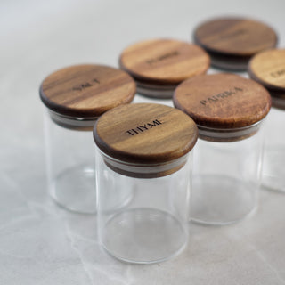 Clear glass spice jars with round wooden lids engraved with names including THYME, PARSLEY, SALT, and PEPPER, arranged on a light gray surface.
