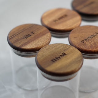 Five clear glass spice jars with round wooden lids engraved with spices like Salt, Pepper, Paprika, and Thyme, arranged on a light gray surface.