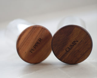 Two round wooden spice grinders labeled 'PEPPER' and 'CUMIN' in dark brown wood, shown close-up on a light background.