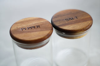 Two clear glass spice jars with round wooden lids engraved with 'PEPPER' and 'SALT', shown on a soft white background.