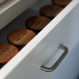 Close-up of a white drawer with a curved metal handle, containing round wooden spice jars labeled OREGANO and others partially visible.