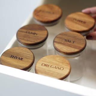 Five small glass spice jars with round wooden lids labeled THYME, OREGANO, SALT, PEPPER, and OREGANO, arranged in a white drawer. A hand is visible touching one lid.