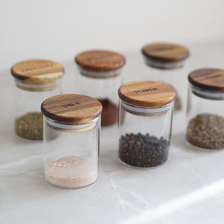 A set of clear glass spice jars with round wooden lids, arranged on a light marble surface. The jars are labeled with engraved wooden lids for 'SALT', 'PEPPER', and other spices.