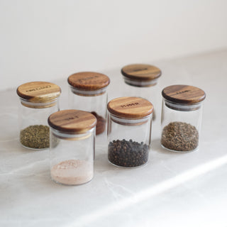 Six small clear glass spice jars with round wooden lids arranged on a light marble surface against a white background. The jars contain various whole and ground spices, and the wooden lids are engraved with spice names including Cumin, Pepper, Salt, and Turmeric.