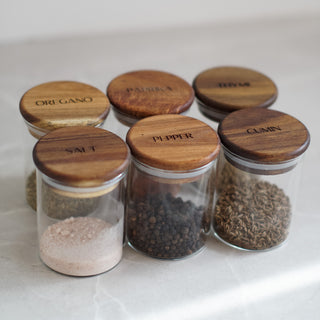 Six clear glass spice jars with round wooden lids labeled Salt, Pepper, Cumin, Oregano, Paprika, and Herbs, arranged on a light marble surface.