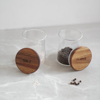 Two clear glass spice jars with round wooden lids engraved with 'SALT' and 'PEPPER', shown on a light marble surface with a few loose peppercorns.