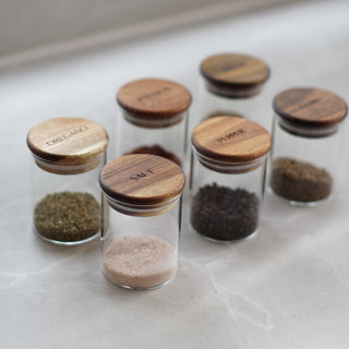 Six small clear glass spice jars with round wooden lids, arranged on a light stone surface. The lids are engraved with spice names including OREGANO, SALT, PEPPER, and others, and the jars contain different ground spices in green, brown, black, and pale pink tones.
