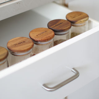 A close-up of a white drawer containing clear glass spice jars with round wooden lids, one of which is engraved with the name 'PAPKA'.