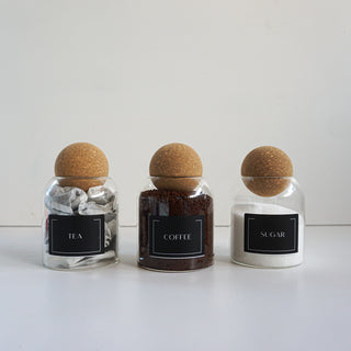 Three clear glass jars with cork stoppers and black labels reading TEA, COFFEE, and SUGAR, arranged on a white surface against a light gray background.