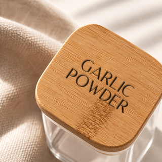 A close-up of a clear glass jar with a square, rounded bamboo lid engraved with the words 'GARLIC POWDER' in dark lettering, resting on a textured beige fabric surface.