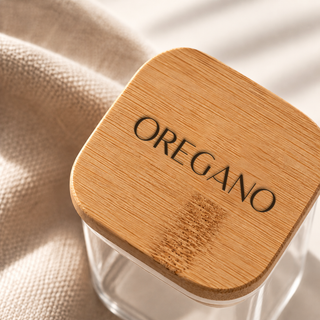 A clear glass jar with a square rounded bamboo lid engraved with the word 'OREGANO', resting on a beige textured fabric surface.