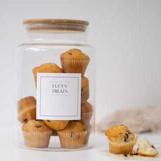 A clear glass jar with a light wood lid, filled with chocolate chip muffins and a white label reading 'LUCY'S TREATS,' on a white surface with one muffin broken in half beside it.
