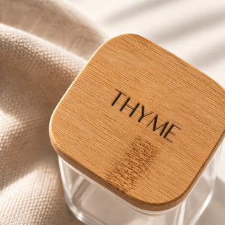 A close-up of a clear glass jar with a square bamboo lid featuring the word 'THYME' engraved on it, resting on a beige textured fabric under soft natural light.