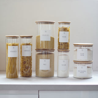 A set of eight airtight glass jars with bamboo lids and white waterproof labels in a kitchen setting.